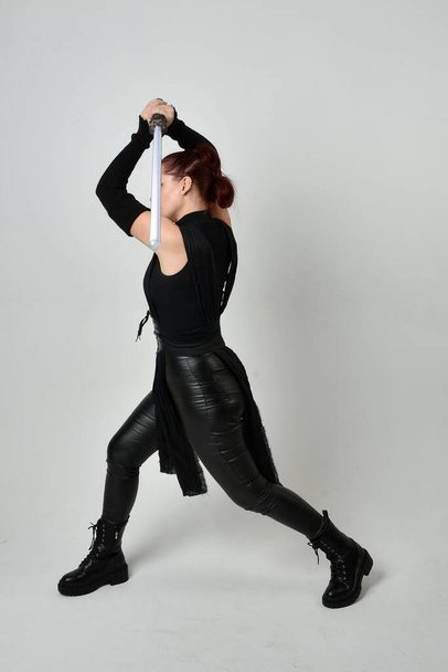 Portrait complet de jolie femme aux cheveux roux portant un costume futuriste noir en cuir de scifi, tenant une arme à épée de sabre laser. poses debout dynamiques avec des mains gestuelles, face à l'arrière sur fond de studio blanc. - Photo, image