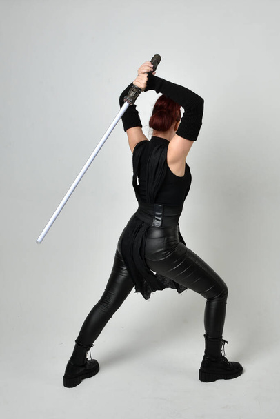 Full length portrait of pretty red haired female model wearing black futuristic scifi leather costume, holding a lightsaber sword weapon. Dynamic standing poses with gestural hands, facing backwards away from  on a white studio background. - Фото, изображение