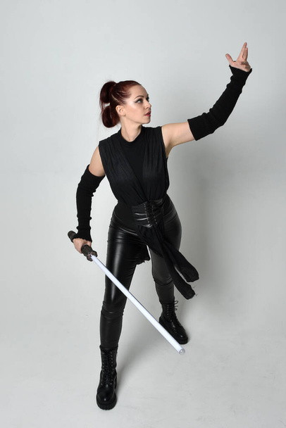 Full length portrait of pretty red haired female model wearing black futuristic scifi leather costume, holding a lightsaber sword weapon. Dynamic standing poses with gestural hands, facing backwards away from  on a white studio background. - Photo, Image