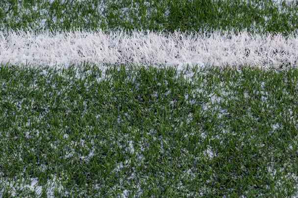 The football field with artificial green grass is covered with a light layer of snow. Early spring. Green grass on the football field is visible from under the snow. Amateur football field. - Photo, Image