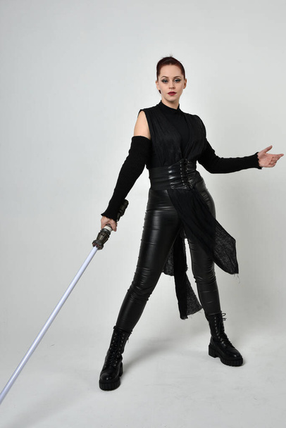 Full length portrait of pretty red haired female model wearing black futuristic scifi leather costume, holding a lightsaber sword weapon. Dynamic standing poses with gestural hands, facing backwards away from  on a white studio background. - Foto, imagen