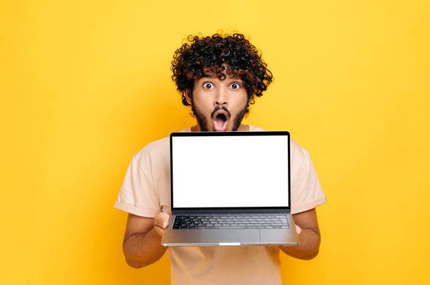 Shocked amazed indian or arabian curly haired guy, holding open laptop, with blank white screen, mockup concept, standing over isolated orange background, looking at camera with open mouth in surprise - Photo, Image