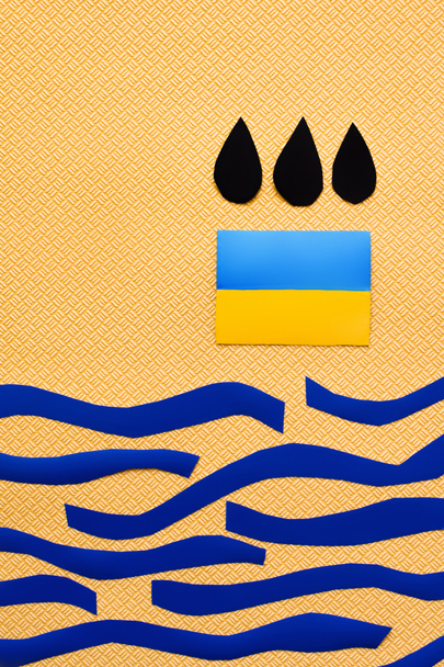 Vue du dessus des gouttes noires au-dessus du drapeau ukrainien et de la mer de papier sur fond jaune texturé  - Photo, image
