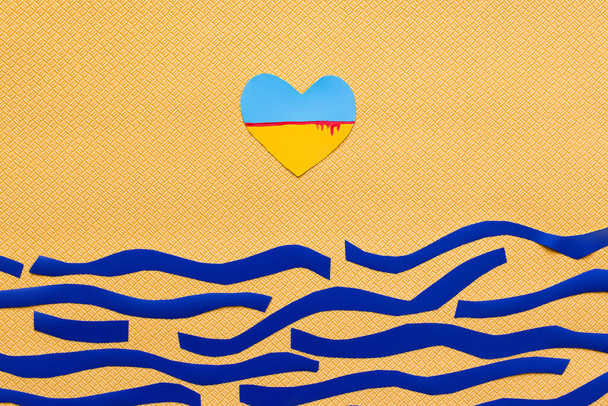 Top view of ukrainian flag with blood in heart shape above paper sea on textured yellow background  - 写真・画像