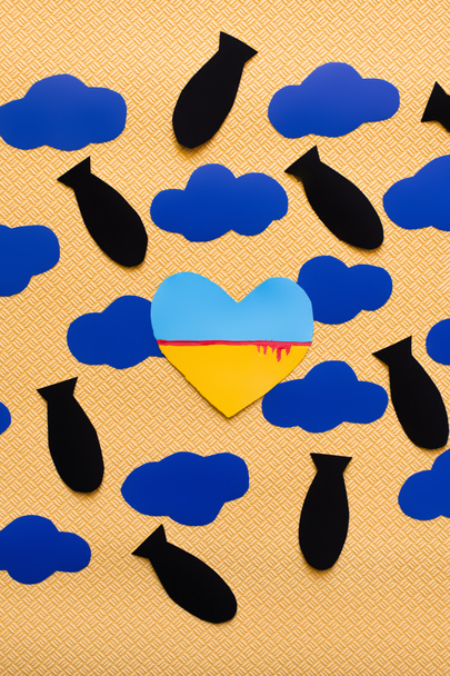Vista superior de la bandera ucraniana en forma de corazón cerca de nubes de cartón y bombas de papel sobre fondo amarillo texturizado  - Foto, Imagen