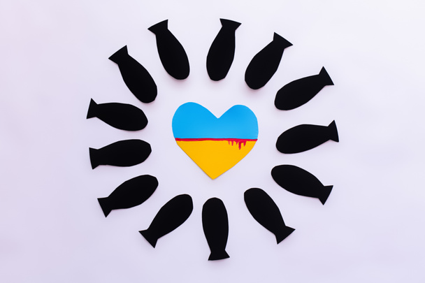 Κάτοψη της ουκρανικής σημαίας σε σχήμα καρδιάς σε πλαίσιο από χάρτινες βόμβες σε λευκό φόντο  - Φωτογραφία, εικόνα