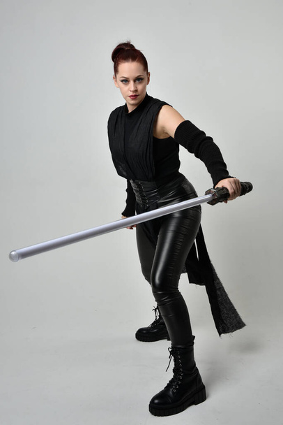 Full length portrait of pretty red haired female model wearing black futuristic scifi leather costume, holding a lightsaber sword weapon. Dynamic standing poses with gestural hands, facing backwards away from  on a white studio background. - Foto, imagen