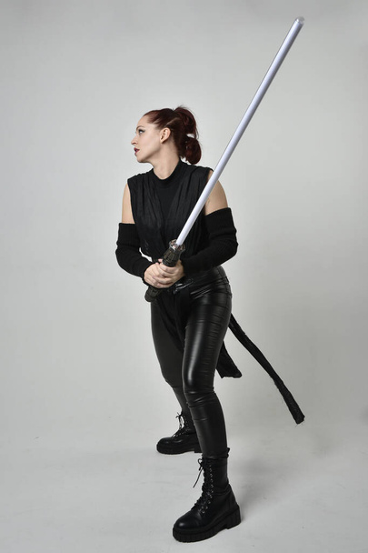 Full length portrait of pretty red haired female model wearing black futuristic scifi leather costume, holding a lightsaber sword weapon. Dynamic standing poses with gestural hands, facing backwards away from  on a white studio background. - Fotografie, Obrázek