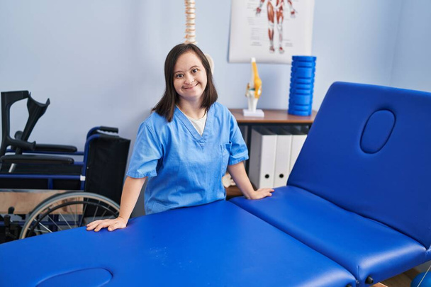 Mulher com síndrome de Down vestindo uniforme de fisioterapia apoiada na mesa de massagem na clínica fisioterapeuta - Foto, Imagem