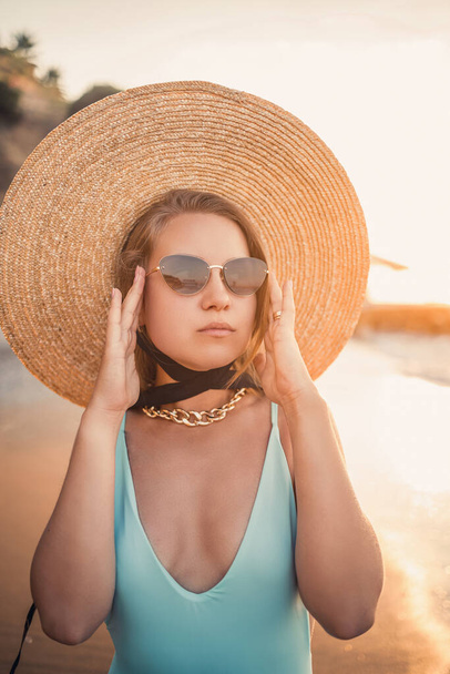 A young tanned woman in a beautiful swimsuit with a straw hat stands and rests on a tropical beach with sand and looks at the sunset and the sea. Selective focus. Vacation concept by the sea - Photo, image