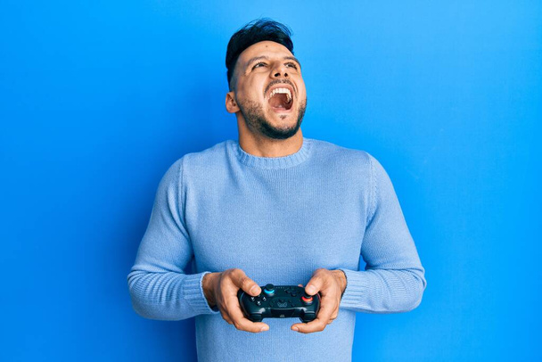 Fiatal arab férfi videojáték holding vezérlő dühös és őrült sikoltozó frusztrált és dühös, kiabál a harag felnézett.  - Fotó, kép