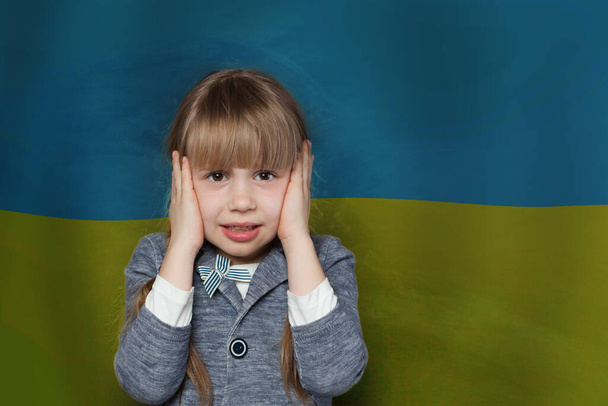 Frightened child girl covers her ears with her hands so as not to hear anything against Ukrainian flag background. Stop war in Ukraine - Photo, Image