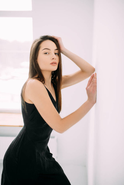 Young slim brunette woman in black dress stands near wall against window. - Photo, Image