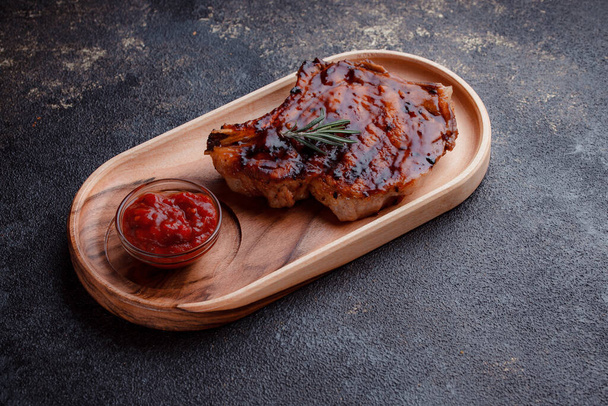 Grilled pork steak in caramelized sauce. Serving meat on eco-friendly wooden utensils. A sprig of fresh rosemary and tomato sauce. - Zdjęcie, obraz