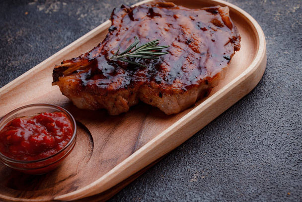 Grilled pork steak in caramelized sauce. Serving meat on eco-friendly wooden utensils. A sprig of fresh rosemary and tomato sauce. - Foto, imagen