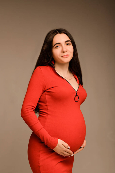 Pregnant woman in a red dress hugs her belly with her hands - Фото, изображение