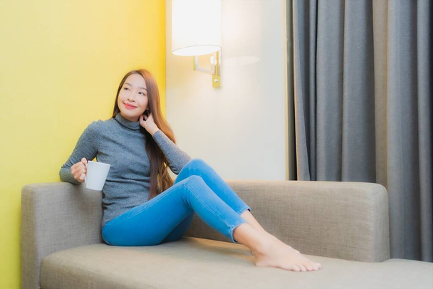 Портрет красива молода азіатка посміхається розслабитися з чашкою кави на дивані в інтер'єрі вітальні
 - Фото, зображення