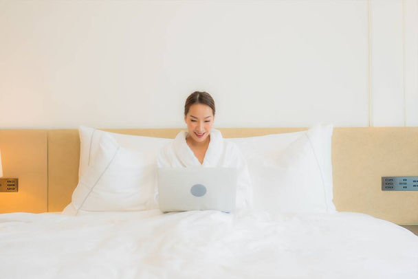 肖像画美しい若いアジアの女性は寝室のインテリアでベッドの上にコンピュータのラップトップを使用します - 写真・画像