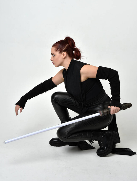 Portrait complet de jolie femme aux cheveux roux portant un costume futuriste noir en cuir de scifi, tenant une arme à épée de sabre laser. pose assise dynamique sur un fond de studio blanc. - Photo, image