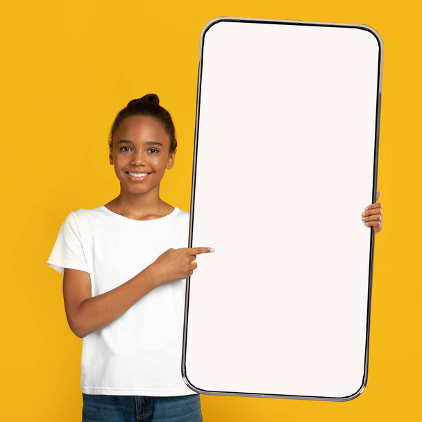 Mooi zwart tienermeisje wijzend naar gigantische smartphone met leeg scherm op oranje achtergrond, mockup voor app-ontwerp - Foto, afbeelding