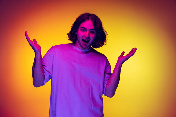 Studio shot of young emotional man, student posing isolated on orange background in purple neon light. Concept of emotions, beauty, fashion - Foto, Imagen