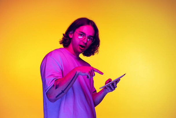 Portrait of young man with long hair using digital gadget isolated on orange background in purple neon light. Concept of emotions, beauty, fashion - Фото, изображение