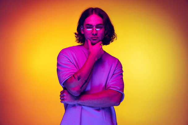 Portrait of young man, student in casual clothes isolated on orange background in purple neon light. Concept of emotions, beauty, fashion - Valokuva, kuva