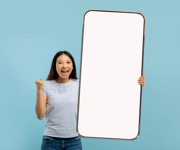 Aufgeregte junge Asiatin hält großes Handy mit leerem Bildschirm, zeigt JA-Geste und bietet Attrappe für Anzeige an - Foto, Bild