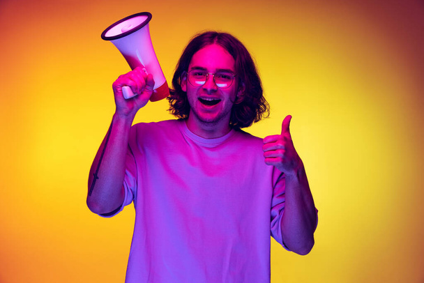 One young handsome man in white t-shirt shouting at megaphone isolated on orange background in neon light. Concept of emotions, fashion, youth - 写真・画像