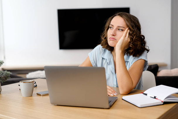 Young caucasian woman looking annoyed and stressed, sitting at the desk, using a laptop, thinking and looking away, feeling tired and bored - Foto, afbeelding