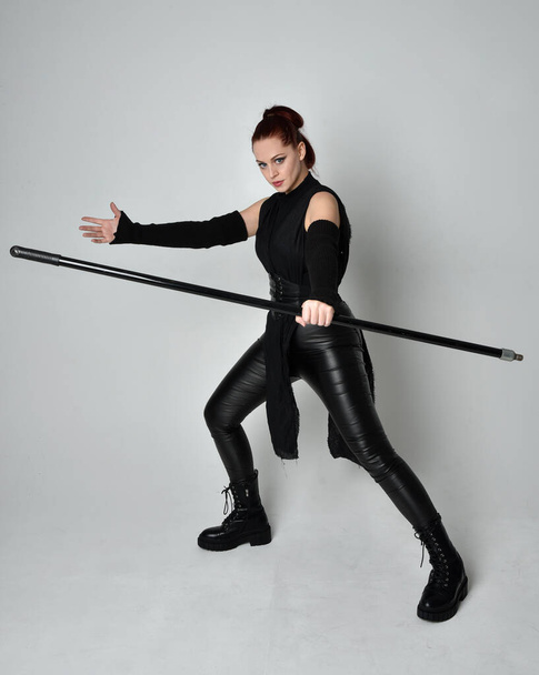 Full length portrait of pretty red haired female model wearing black futuristic scifi leather costume, holding a staff spear weapon. Dynamic standing pose on a white studio background. - Fotografie, Obrázek