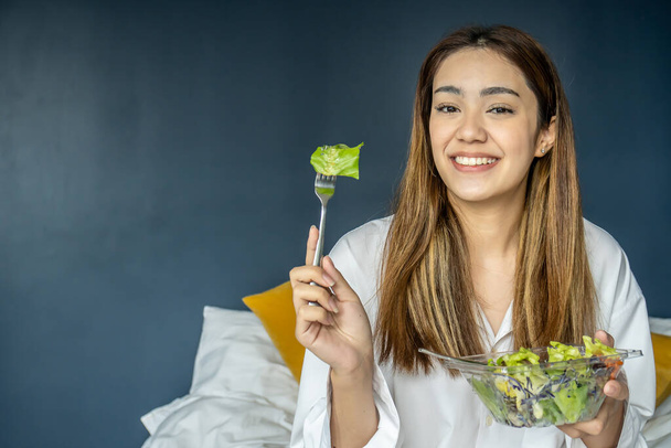 Prachtige jonge vrouw die op bed zit en groentesalade eet, evenwichtige rauwe voeding. Positief meisje met veganistische lunch, zorgen voor haar welzijn - Foto, afbeelding