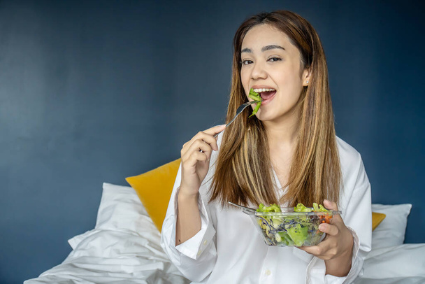 Gyönyörű fiatal nő ül az ágyon, zöldségsalátát eszik, kiegyensúlyozott nyers étel diéta. Pozitív lány vegetáriánus ebéd, gondoskodás a jólét - Fotó, kép