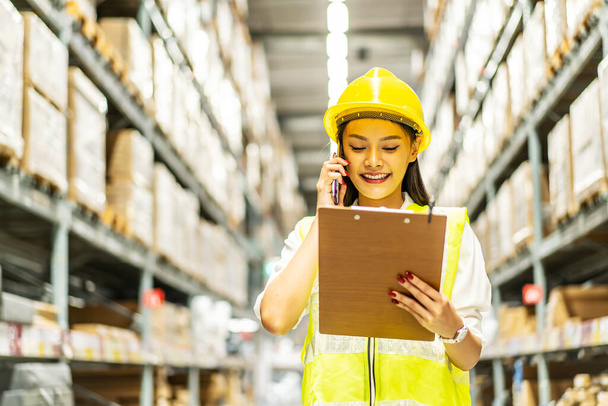 Asian woman in helmet and uniform talks on mobile phone discuss amount of stock product inventory on shelf at distribution warehouse factory. logistic business shipping and delivery service - Zdjęcie, obraz