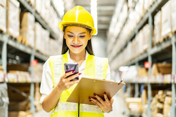 Young asian woman worker uses the phone for business contacts and holding clipboard to checking inventory in the warehouse - Photo, Image