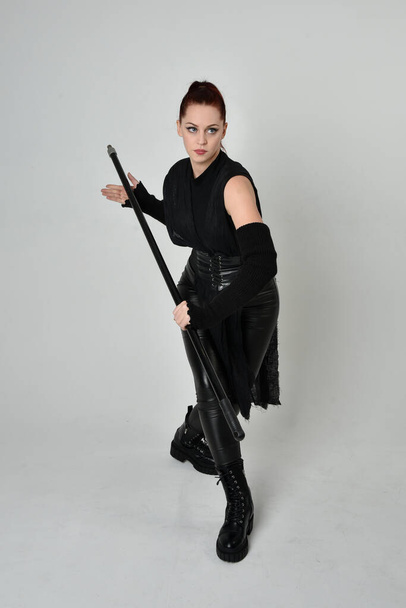 Retrato de comprimento total do modelo feminino ruivo bonito vestindo traje de couro scifi futurista preto, segurando uma arma de lança pessoal. Posição de pé dinâmica em um fundo de estúdio branco. - Foto, Imagem
