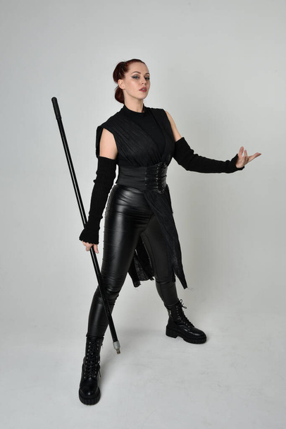 Full length portrait of pretty red haired female model wearing black futuristic scifi leather costume, holding a staff spear weapon. Dynamic standing pose on a white studio background. - Photo, Image