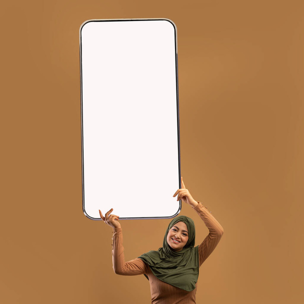 Cheerful young Arab woman in hijab holding huge cellphone with blank screen, offering newest mobile app, mockup - Photo, image