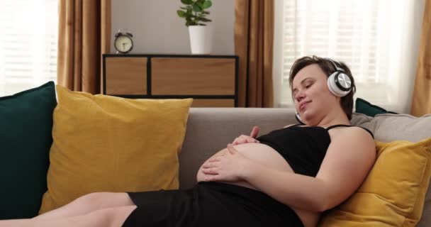 お腹にヘッドフォンをしている妊婦さん。白人妊娠中の女性でベッドの上にヘッドフォンを持っています彼女の裸の腹とストロークそれの間に赤ちゃん聞く音楽 - 映像、動画