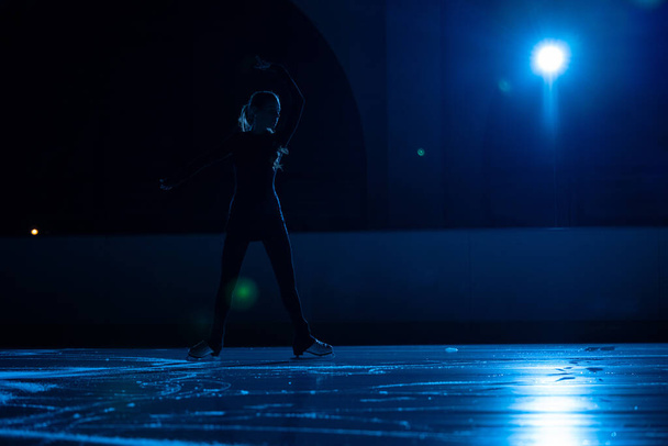 Young female figure skater is performing womans single skating choreography on ice rink. Dark silhouette of woman practicing skills on ice arena against background of soft blue light and spotlights. - Zdjęcie, obraz