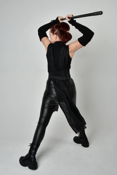 Portret pięknej rudej modelki w czarnym futurystycznym skórzanym kostiumie, trzymającej broń z włóczni. Dynamiczna pozycja stojąca do tyłu, do tyłu, białe tło studia. - Zdjęcie, obraz