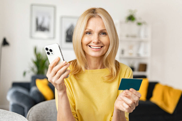 Middle-aged caucasian woman consumer holding credit card and smartphone sitting on sofa at home making online banking transaction - Foto, Bild
