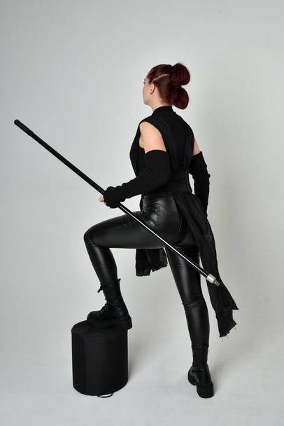 Ganztägiges Porträt eines hübschen rothaarigen weiblichen Modells im schwarzen futuristischen Scifi-Lederkostüm, das eine Stabspeerwaffe in der Hand hält. Dynamische Haltung im Stehen mit dem Gesicht nach hinten, weißer Studiohintergrund. - Foto, Bild