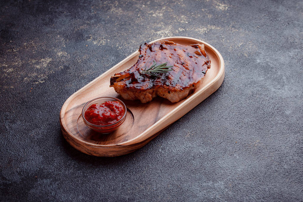 Flat lay of grilled pork steak in caramelized sauce. Serving meat on eco-friendly wooden utensils. A sprig of fresh rosemary and tomato sauce. - Zdjęcie, obraz