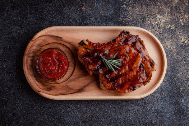 Flat lay of grilled pork steak in caramelized sauce. Serving meat on eco-friendly wooden utensils. A sprig of fresh rosemary and tomato sauce. - Photo, Image