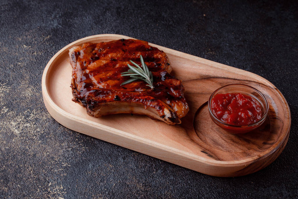 Flat lay of grilled pork steak in caramelized sauce. Serving meat on eco-friendly wooden utensils. A sprig of fresh rosemary and tomato sauce. - Photo, Image