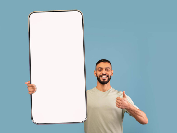 Sonriente joven árabe demostrando un teléfono inteligente gigante con maqueta, mostrando pulgares hacia arriba, promoviendo aplicación o sitio web - Foto, Imagen