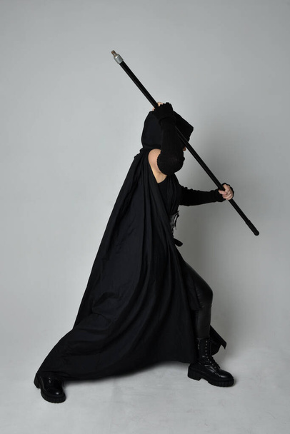 Ολόσωμο πορτραίτο μιας όμορφης κοκκινομάλλας που φορούσε μαύρη φουτουριστική στολή επιστημονικής φαντασίας, μαύρο μανδύα που κρατούσε ένα ψαροντούφεκο. Δυναμική στάση σε λευκό φόντο στούντιο. - Φωτογραφία, εικόνα