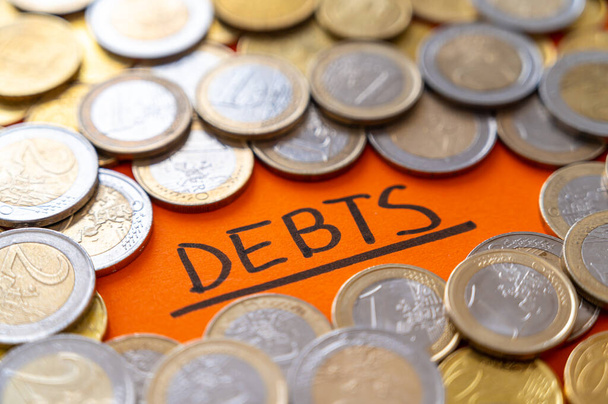Text "Schulden", schwarz auf orangefarbener Fläche geschrieben, mit Münzen drumherum. Steigende Inflation, Schulden und Wirtschaftskrise.  - Foto, Bild