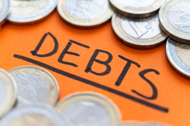 Text "dluhy", napsaný černě na oranžovém povrchu, s mincemi kolem sebe. Rostoucí inflace, zadlužení a hospodářská krize.  - Fotografie, Obrázek